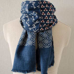 May include: A blue scarf with white dragonfly, geometric, and triangle patterns. The scarf is draped over a mannequin. The scarf has a fringed edge and is made of a soft fabric. The scarf is a fashion accessory.