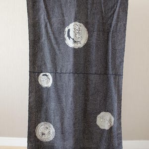 May include: Dark gray fabric with a geometric pattern and circular illustrations. The fabric is hanging vertically, showcasing a design with vertical lines and square accents. The circular illustrations feature detailed drawings in white.
