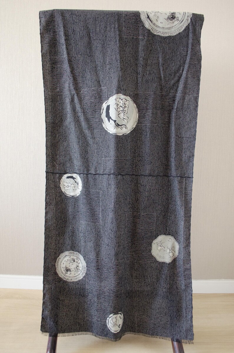 May include: Dark gray fabric with a geometric pattern and circular illustrations. The fabric is hanging vertically, showcasing a design with vertical lines and square accents. The circular illustrations feature detailed drawings in white.