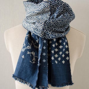 May include: A blue and white patterned scarf displayed on a mannequin. The scarf features various designs, including wave patterns, geometric shapes, dragonflies, and star-like motifs. The edges are fringed.