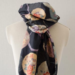 May include: A dark blue scarf with a repeating pattern of floral medallions in shades of red, yellow, and purple. The scarf is draped over a mannequin, showcasing its design and texture. The edges have a frayed finish.
