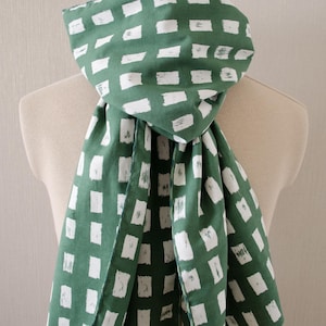 May include: A green scarf with a white rectangular pattern is draped over a mannequin. The scarf is made of a soft fabric and has a repeating design of white rectangles against a green background. The scarf is folded and draped to showcase the pattern.