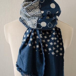 May include: A blue and white patterned scarf draped over a mannequin. The scarf features wave, dot, and crosshatch designs. The scarf has a fringed edge and is made of a soft fabric.