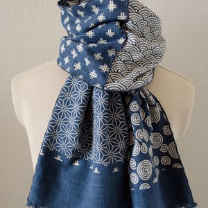 May include: A blue and white patterned scarf with various geometric and wave designs. The scarf is draped over a mannequin, showcasing its fringed edges and intricate patterns. The scarf's design includes starbursts, cross-hatch, and spiral patterns.