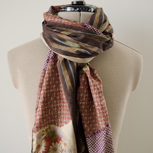 May include: A rectangular scarf featuring a patchwork design with brown, red, and purple tones. The fabric displays a repeating geometric pattern, vertical stripes, and a floral print with white daisies and red flowers on a light background.