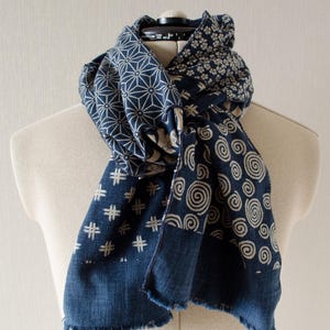 May include: A blue and white patterned scarf draped over a mannequin. The scarf features various geometric and floral designs, including star-like shapes, small flowers, and spiral patterns. The edges have a frayed, unfinished look.