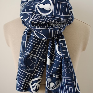 May include: A navy blue scarf featuring a white grid pattern with various botanical and geometric illustrations, including pears, flowers, and abstract shapes. The scarf is draped in a loop, displaying a repeating print across the fabric surface.