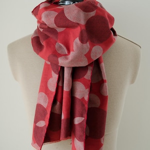 May include: A red scarf featuring a repeating pattern of large, overlapping circles in varying shades of dark red and light pink. The scarf has a textured, woven appearance with raw, unfinished edges and is draped around a neutral-coloured mannequin.