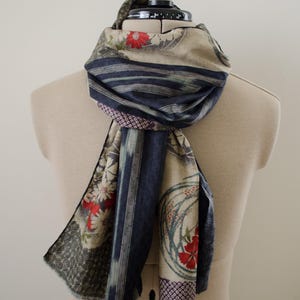 May include: A scarf featuring a multi-patterned design with beige, dark blue, and red floral motifs. The fabric displays a combination of circular floral arrangements, geometric patterns, and vertical striped sections in a layered, draped style.