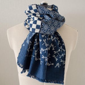 Puede incluir: Bufanda estampada en azul y blanco sobre un maniquí. La bufanda presenta tres diseños distintos: un tablero de ajedrez, un motivo geométrico de estrella y un motivo de libélulas. La bufanda tiene un borde con flecos.