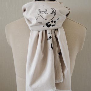 May include: Beige scarf with a black cat illustration. The scarf is draped over a mannequin and features a playful cat design on the front. The scarf has a fringed edge.