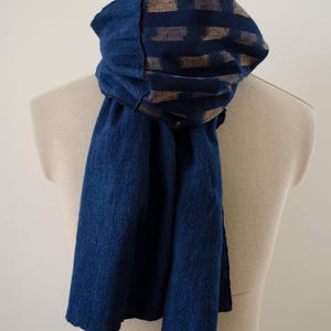 Puede incluir: Una bufanda azul profundo con un patrón de diamantes tejido y texturizado en toda la superficie. La sección superior presenta una banda horizontal de bloques rectangulares azul oscuro y beige. La bufanda está drapeada de forma holgada.