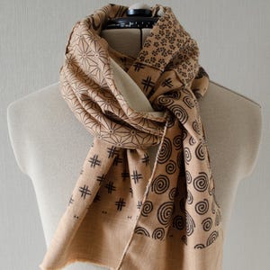 May include: A tan scarf with various black geometric patterns, including spirals, hash marks, and starburst shapes. The scarf is draped over a mannequin, showcasing its design and texture. The scarf has a fringed edge.