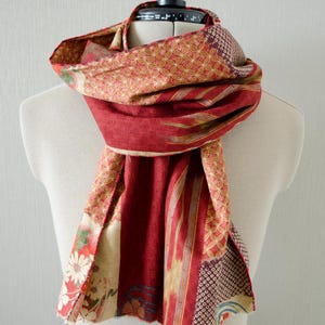 May include: A red and patterned scarf draped over a mannequin. The scarf features a mix of floral, geometric, and striped patterns in shades of red, gold, and cream. The scarf is made of a soft, flowing fabric.