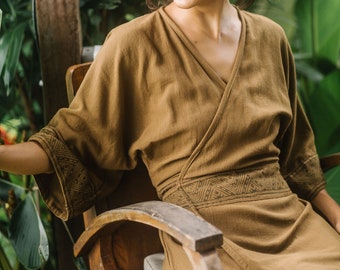 Puede incluir: Una mujer con un vestido marrón de envoltura con un patrón geométrico se sienta en una silla de madera. Ella sonríe y mira hacia un lado. El fondo es un exuberante jardín tropical verde.
