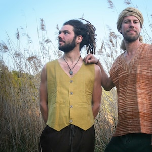May include: Two people wearing sleeveless vests stand in a field of tall grass. The person on the left is wearing a yellow vest with buttons and a necklace. The person on the right is wearing a brown and orange striped vest.