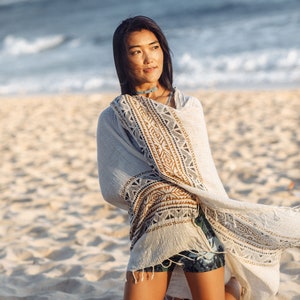 May include: A woman wearing a white shawl with a geometric pattern in brown and black, kneeling on a sandy beach. The shawl is draped over her shoulders and billowing in the wind. She is looking off to the side with a thoughtful expression.