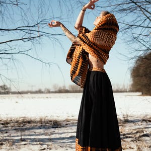 May include: A woman wearing a long black skirt with an orange and brown patterned trim. She is wearing a patterned orange and brown shawl and has her arms raised in a yoga pose. The background is a snowy field with bare trees.