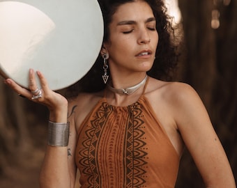 May include: A woman holding a Remo tambourine. She wears a halter top with a brown tribal pattern, silver jewelry, and a silver cuff bracelet. The background is blurred with natural light.
