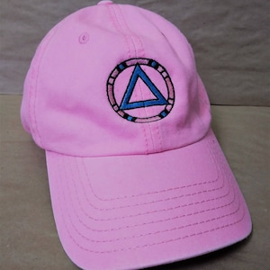 May include: A pink baseball cap with a curved brim. The cap features an embroidered emblem of a blue triangle within a circle, with black and pink accents. The cap is made of a soft fabric and has a structured crown.