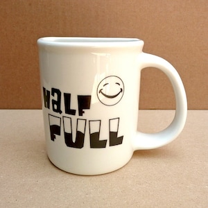 Puede incluir: Taza de cerámica blanca con texto negro que dice "half full" y una cara sonriente negra.