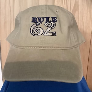 May include: A khaki baseball cap with the text "RULE 62" embroidered in black and white.
