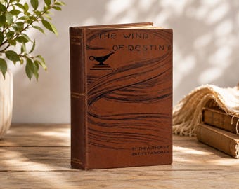 May include: A brown hardcover book titled The Wind of Destiny by the author of But Yet a Woman. The front cover features black line art of a genie lamp and swirling wind patterns. The title and author text are printed in a stylized black font.