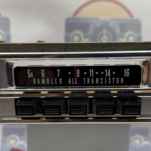 May include: A vintage Rambler All Transistor car radio featuring a chrome faceplate, two black circular knobs on the sides, and five black rectangular push buttons below a black display panel with white frequency numbers ranging from 5 to 16.
