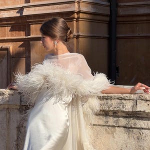 May include: A floor-length cream-colored satin slip dress featuring a sheer organza overlay. The garment is adorned with a thick, voluminous white feather trim along the neckline and down the length of the sheer cape-like overlay.