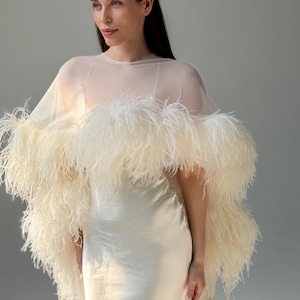 May include: A cream-colored floor-length slip dress paired with a sheer, lightweight capelet. The capelet features a wide, voluminous trim of long, soft, off-white ostrich feathers that drape over the shoulders and extend down the arms.