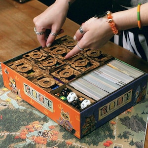 May include: A Root board game organizer box featuring orange and blue sides with owl illustrations and the text Root. The box contains laser-cut wooden inserts, stacks of playing cards, and several six-sided dice with various symbols and numbers.