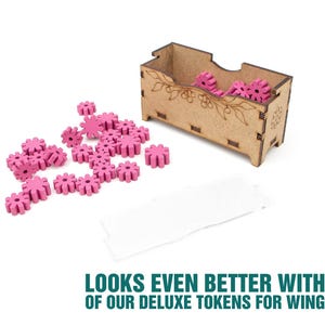 May include: A rectangular wooden storage box with laser-etched floral details, containing several small, bright pink, gear-shaped tokens. Additional pink gear-shaped tokens are scattered on the surface next to the box, alongside a blank white rectangular card.