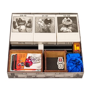 Op de afbeelding: Een bordspel organizer doos met drie witte kaartvakken gelabeld 09-19, 31-42 en 55-66. Het bevat houten bakjes met een kaartspel met een Double Detector illustratie, een kleine witte karakter-token, gele blokjes en een blauw trekkoordzakje.
