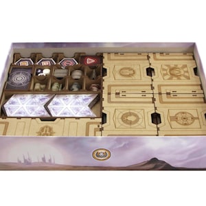 May include: A board game box organiser made of light brown laser-cut wood, featuring multiple compartments for game components. The box interior contains various tokens, cards, and wooden dividers with etched geometric patterns and symbols.