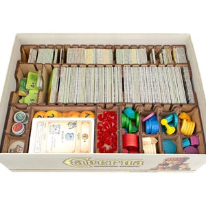 May include: A board game organizer box made of light brown wood, featuring multiple compartments filled with stacks of rectangular game cards, small circular tokens in various colors, red translucent gems, and wooden shapes.