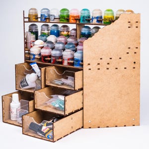 May include: A light brown wooden craft organiser featuring multiple shelves filled with various small paint pots and several drawers containing glue bottles, small metal tools, and plastic bags with supplies. The unit has a tall side panel.