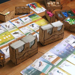May include: A board game setup featuring laser-cut wooden organizers holding card decks, small wooden bird tokens, and colorful egg tokens. The game includes illustrated bird cards, player boards with green and blue gradients, and dice.