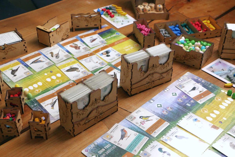 May include: A board game setup featuring laser-cut wooden organizers holding card decks, small wooden bird tokens, and colorful egg tokens. The game includes illustrated bird cards, player boards with green and blue gradients, and dice.
