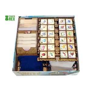 Puede incluir: Un organizador de juegos de mesa de madera para Stardew Valley que contiene dos tarjeteros, una cuadrícula de fichas de objetos cuadradas con ilustraciones coloridas, una bolsa de tela azul y una bolsa de tela beige en una caja.
