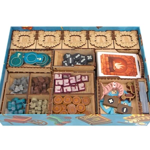 May include: A wooden board game organizer insert inside a blue box, featuring multiple compartments filled with game components including teal circular tokens, black markers, a deck of cards, purple blocks, brown coins, and various wooden shapes.