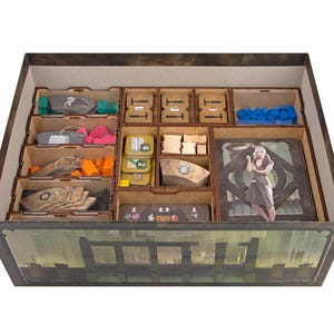 Pode incluir: Uma caixa organizadora de madeira para jogos de tabuleiro com múltiplos compartimentos contendo vários componentes de jogo, incluindo fichas azuis, peças de madeira, baralhos de cartas e um suporte de personagem com uma pessoa de vestido cinzento.
