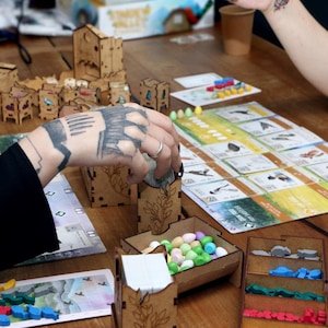 May include: A tabletop board game setup featuring laser-cut wooden organizers, card holders, and trays filled with small colorful egg-shaped tokens. The game board displays various bird illustrations, while players interact with the wooden components.