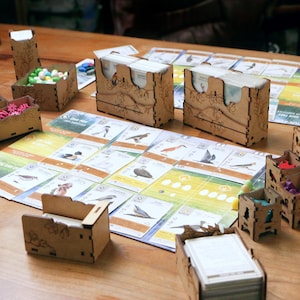 May include: A board game setup featuring laser-cut wooden organizers and card holders. The game board displays various bird illustrations and text. Small wooden containers hold colorful tokens, while stacks of cards are placed in wooden boxes.