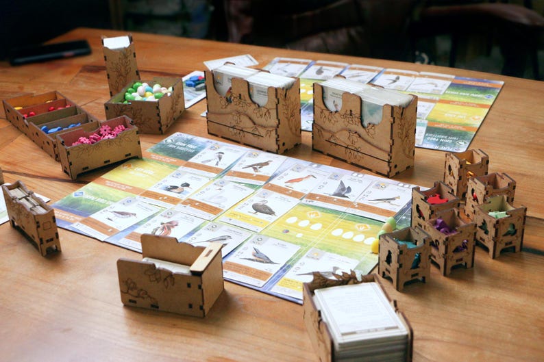 May include: A board game setup featuring laser-cut wooden organizers and card holders. The game board displays various bird illustrations and text. Small wooden containers hold colorful tokens, while stacks of cards are placed in wooden boxes.