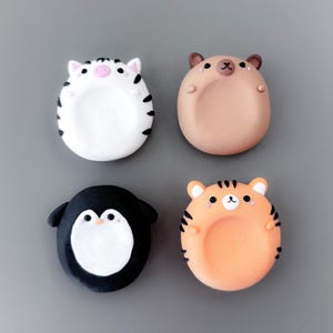 May include: Four colorful, cartoon animal-shaped magnets. The set includes a zebra, a bear, a penguin, and a tiger. Each magnet has a rounded shape with a recessed center. The zebra is white with black stripes, the bear is tan, the penguin is black and white, and the tiger is orange with black stripes.