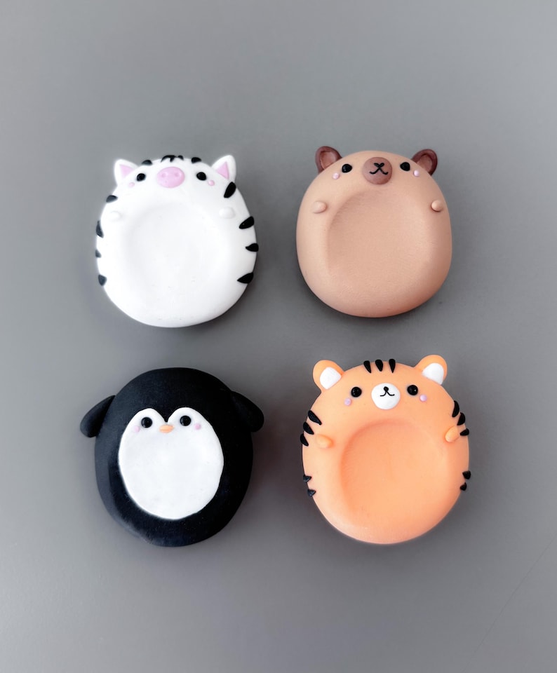 May include: Four colorful, cartoon animal-shaped magnets. The set includes a zebra, a bear, a penguin, and a tiger. Each magnet has a rounded shape with a recessed center. The zebra is white with black stripes, the bear is tan, the penguin is black and white, and the tiger is orange with black stripes.