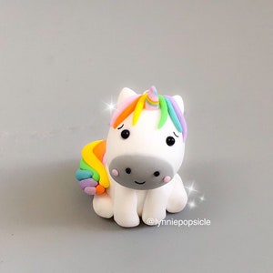 Könnte beinhalten: Eine kleine, weiße Einhornfigur mit einer Regenbogenmähne und einem Regenbogen-Schwanz. Das Einhorn hat eine graue Nase und schwarze Augen. Die Figur sitzt auf einer grauen Oberfläche. @lynniepopsicle