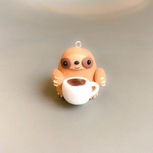May include: A brown sloth figurine holding a white coffee cup with brown liquid inside. The sloth has black eyes and a small smile.