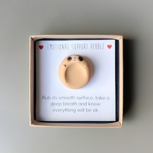May include: A tan-colored emotional support pebble shaped like a sloth, presented in a small box. The text "EMOTIONAL SUPPORT PEBBLE" is above the pebble. The text below reads "Rub its smooth surface, take a deep breath and know everything will be ok."