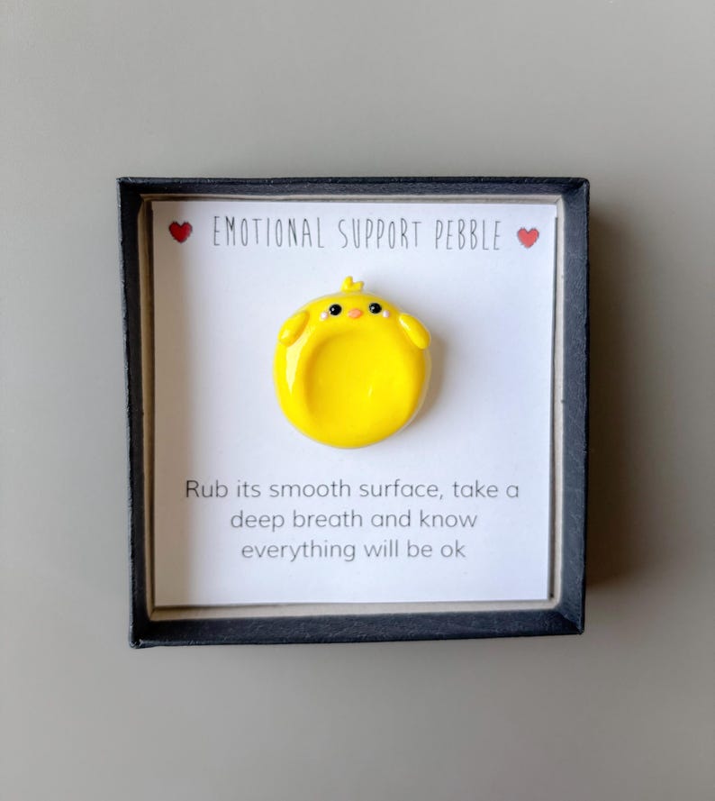 May include: A yellow, chick-shaped emotional support pebble in a black frame. The text "EMOTIONAL SUPPORT PEBBLE" is above the pebble, with two red hearts. Below the pebble is the text: "Rub its smooth surface, take a deep breath and know everything will be ok."
